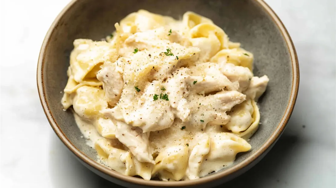 A bowl of Chicken Tortellini Alfredo garnished with black pepper and fresh herbs.